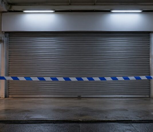 Shuttered front of a neighbourhood minimart at night with blue and white cordon tape across it