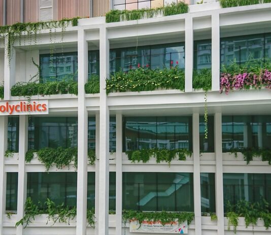 Exterior of Punggol Polyclinic at Oasis Terraces in Singapore, with the orange SingHealth Polyclinics sign on its green, plant-covered facade (Photo: Deoma12 / Wikimedia Commons, CC BY-SA 4.0).